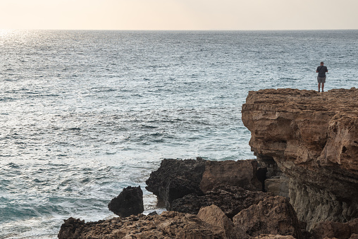 Unrecognized person standing at the edge of rocky coast sightseeing enjoying sunset. Cape Greco Cyprus