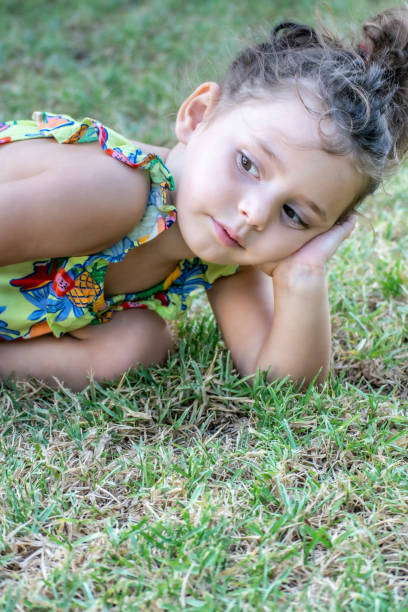 A little girl rests pleasantly in the park, holding her head with her hand while enjoying the warm summer sun and the freshness of the grass.