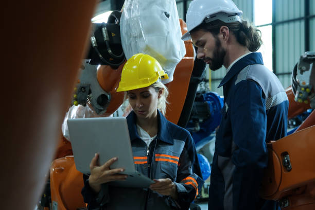 istock Young female robotic engineer wearing safety helmet using laptop with male worker for inspection automation robot arm in factory. 2017363929