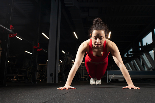 young athletic girl in red sportswear is doing push-ups from the floor in a black gym, an attractive woman is training in a dark fitness room