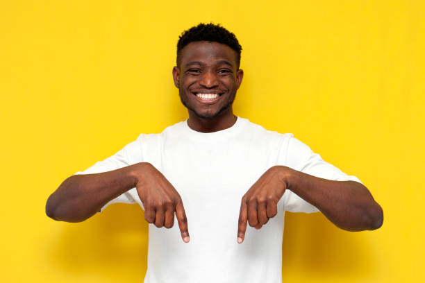 joyful african american man in white t-shirt shows his hands down on yellow isolated background joyful african american man in white t-shirt shows his hands down on yellow isolated background, young guy advertises copy space from below and points man hand down stock pictures, royalty-free photos & images