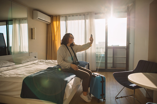 Woman with suitcase in a hotel room communicating with friends or family through smartphone. Summer travel and tourism concept.