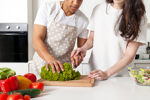 multiracial young couple preparing veggie vegetable and greens salad in white modern kitchen, african american man in apron cuts greens, wife helps and teaches husband to cook