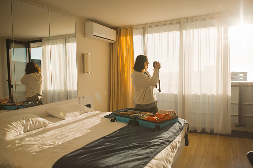 Woman with luggage in a hotel room taking photos through window. Summer travel and tourism concept.