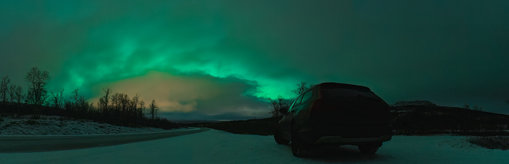 Scenic frosty night panorama of dark car on mountain road in front of Northern green lights shine over mountains in Sweden, Lapland. Night photo, Aurora Borealis, rear side photo