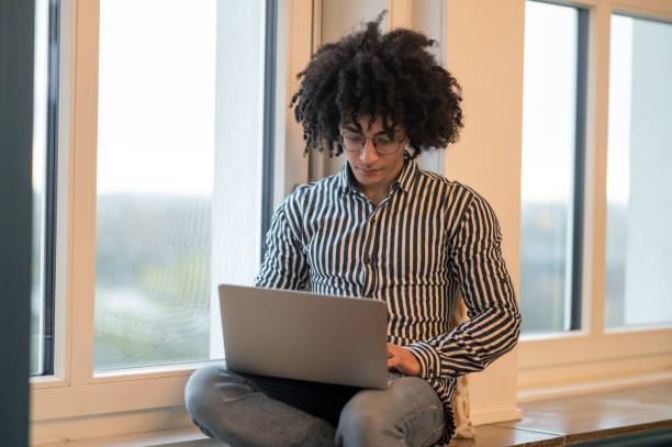 istock Curly-haired man sitting on a window-sill and working on laptop 2017429057