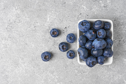 Fresh blueberries in a small rectangular white bowl with scattering of berries on gray concrete background. Top view with copy space for text. Organic berries, healthy food, wild berries