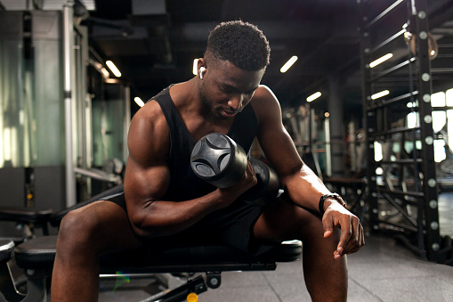 young athletic african american man trains in dark gym, athletic guy lifts heavy dumbbells in fitness club