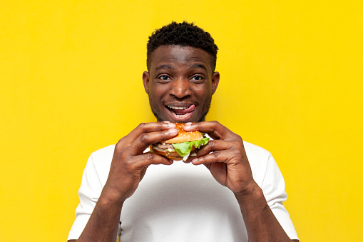 african american man in white t-shirt holds big burger and licks his lips on yellow isolated background, young guy eats fast food and smiles