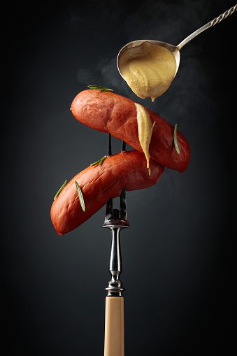 Boiled sausages with rosemary and mustard on a fork. Hot sausages with smoke on a black background.