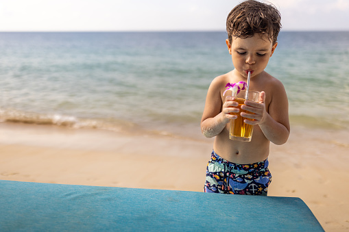 Cute little boy drinking apple juice on the beach by the sea, he is on vacation in Thailand.