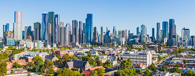 Aerial wide angle panoramic view Melbourne CBD with impressive clusters of high-rise buildings and architecture, varied eras of North Melbourne buildings and architecture, tree-lined streets in foreground. Multiple logos, none dominant.