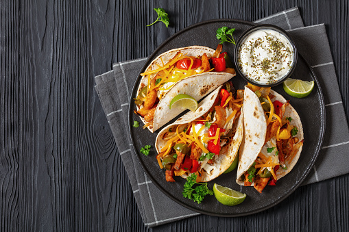 baked tex-mex chicken fajitas with mixed sweet pepper, onion, sour cream, shredded cheese and white corn tortillas on black plate on black wooden table, horizontal view,  flat lay, free space