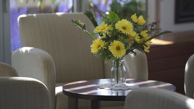 A bright beautiful bouquet of yellow flowers on a coffee table, surrounded by comfortable fabric armchairs lit by the afternoon springtime sun streaming through the windows.