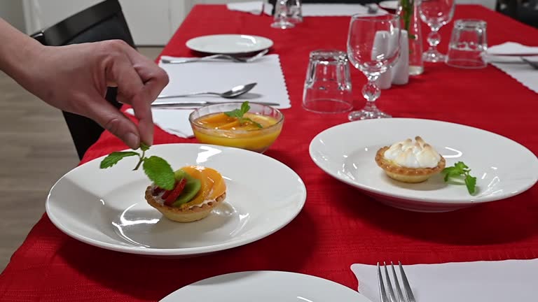 A waiter places a fresh mint leaf as a garnish on a delicious sweet fruit tart filled with cream and summer fruits, on a table in a hotel restaurant.