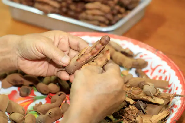 Photo of A close-up of an elderly Asian man preparing soggy tamarind by cracking ripe, sour tamarind shells on a wooden table at home.