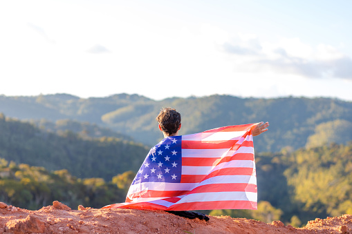 A man seated atop a mountain, proudly draped in the US flag, surrounded by a mountainous landscape. Ideal for campaigns celebrating unity during sports events or in commemoration of July 4th, Independence Day in the United States.