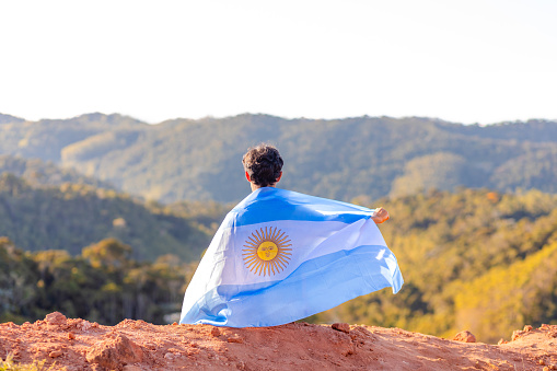 A man seated atop a mountain, proudly holding the Argentine flag, surrounded by a mountainous landscape. Ideal for campaigns celebrating national pride during sports events or in commemoration of Argentina's Independence Day
