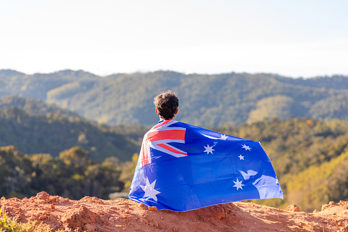 A man seated atop a mountain, proudly holding the Australian flag, surrounded by a mountainous landscape. Ideal for campaigns celebrating national pride during sports events or in commemoration of significant Australian national holidays