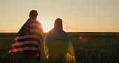 Couple with the flags of Ukraine and the USA stand side by side and look at the sunset over a field of wheat