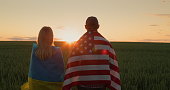 A man and a woman with the flags of Ukraine and the USA stand side by side and look at the sunrise over a field of wheat