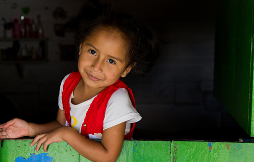 indigenous girl standing in the window of her house looking at the camera and smiling slightly