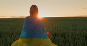 A woman with the flag of Ukraine on her shoulders looks at the sunrise over a field of wheat. Stand with Ukraine concept