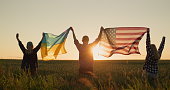 A group of friends raises the flag of the United States and the flag of Ukraine over their heads. Standing against the backdrop of a field of wheat where the sun sets