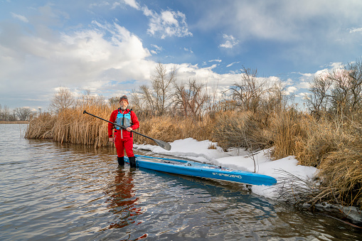 Fort Collins, CO, USA - February 15, 2024. Senior paddler wearing life jacket and drysuit is standing in water next to his Starboard stand up paddleboard on lake in Colorado in winter scenery