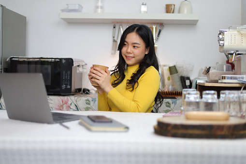 Young Asian woman drinking coffee working with laptop in kitchen at home.