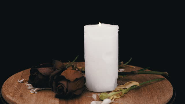 Burning White Candle along with Two Withered Dry Roses on a Black Background