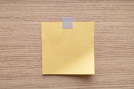 A single yellow sticky note affixed to a wooden textured surface with a tape.