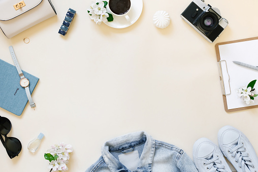 Planning a summer trip. A tablet with a blank sheet of paper, a retro camera, white apple blossoms, a watch, women's clothing and a cup of coffee on a beige background with a copy space. The workplace of a travel blogger