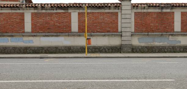 Orange waste bin hanged on yellow pole on concrete sidewalk. Brick and cement wall with erased graffiti on behind, street in front.