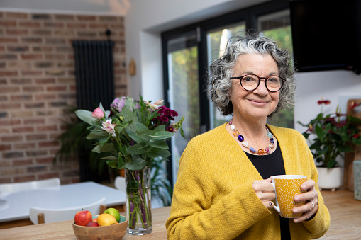 Portrait of a mature adult holding a coffee mug at home. She is wearing a yellow cardigan and spectacles and is looking at the camera.