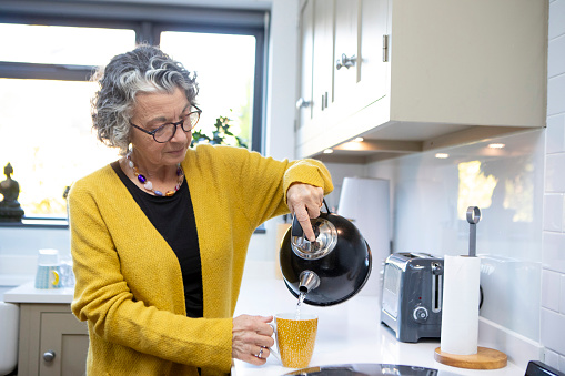 A mature adult pouring water from a kettle into a yellow mug. She is wearing a yellow cardigan and spectacles.