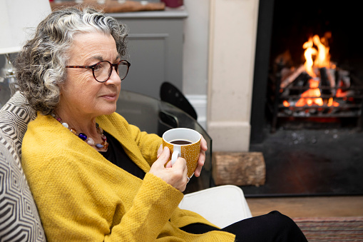 A mature adult sitting by a fireplace in a living room at home. She is wearing a yellow cardigan and spectacles.