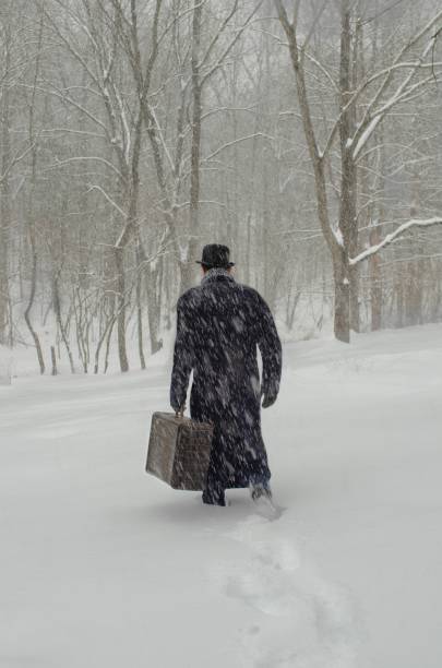 Retro Man walking away in snow carrying vintage suitase wearing long coat boler hat stock photo