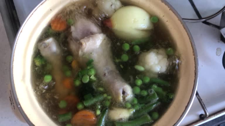 Horizontal straight from above video of a chicken soup boiling in a yellow pan on an old fashioned stove. Children legs, carrots, green peace, onion are visible. A woman’s hand mixes it with a silver tablespoon and covers with a cap.