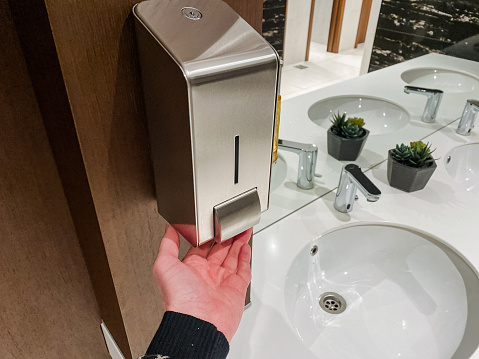 A person is operating a soap dispenser in a bathroom of a building with hardwood flooring, wood cabinetry, and wood stain.