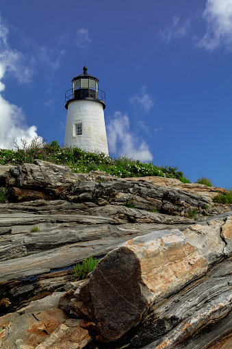 Pemaquid Point Lighthouse in Bristol Maine sits atop rugged rock layers on a bright summer day dotted with puffy clouds