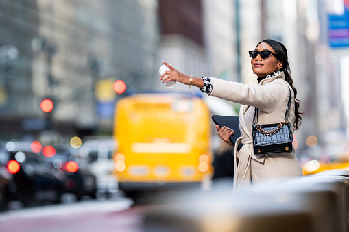 A casually dressed female entrepreneur seen in a bright coat holding a laptop in one hand and a cup of coffee in the other while stopping a taxi in New York City during one busy work day.