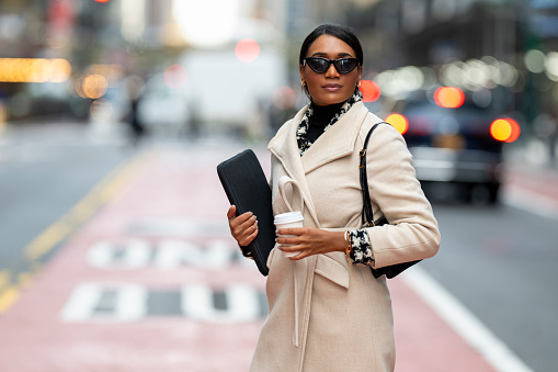 A beautiful female entrepreneur wearing bright coat and sunglasses seen on the street of Manhattan while holding a laptop and a coffee cup and waiting for an Uber after finishing work.