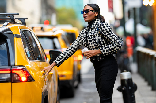 A beautiful female entrepreneur with black sunglasses in a plaid jacket and with a purse seen entering a yellow cab in Manhattan during one busy day.