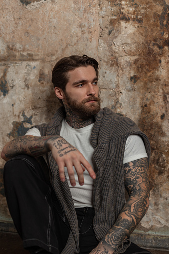 Cool style brutal man with a hairstyle and beard with a tattoo in fashion clothes with a T-shirt and sweater sits near a vintage grunge wall