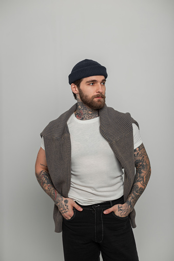 Handsome fashionable hipster man with a beard with tattoos in a fashion white T-shirt with a knitted sweater and hat stands on a white background in the studio
