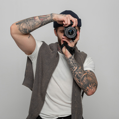 Handsome creative hipster man with tattoos in a hat in a fashion knitted sweater takes pictures with a mirrorless photo camera in the studio