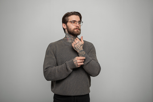 Stylish handsome successful hipster man with a beard and hairstyle with fashionable glasses in a knitted vintage sweater on a white background in the studio