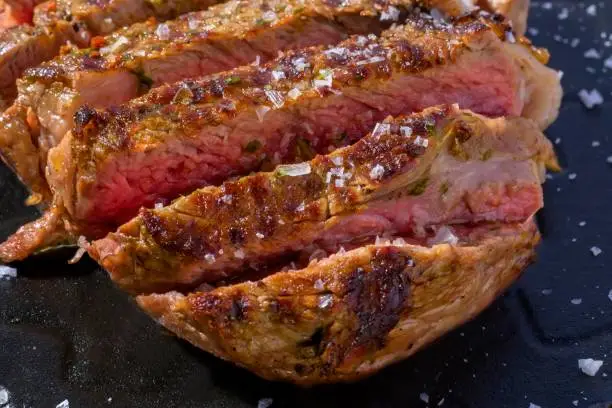 Close-up of a cut of beef on a black plate with salt. The cut is approximately 2 cm thick and has a slightly marbled texture. The meat is cooked to medium rare, with a pink color in the center and a golden crust on the outside.