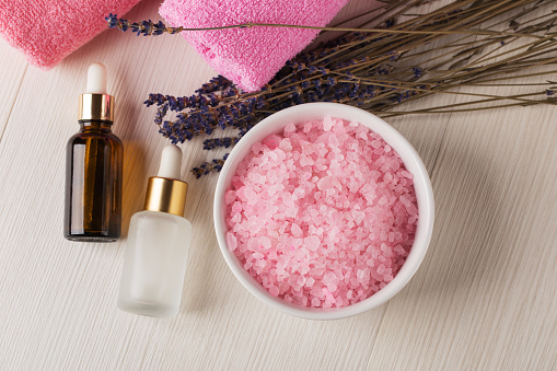 Bowl of spa sea salt with essential oils, dropper bottles with lavender cosmetic oil and face serum, dry lavender flowers on white wooden table. Herbal cosmetics and aromatherapy concept. Lavender beauty products. Top view.